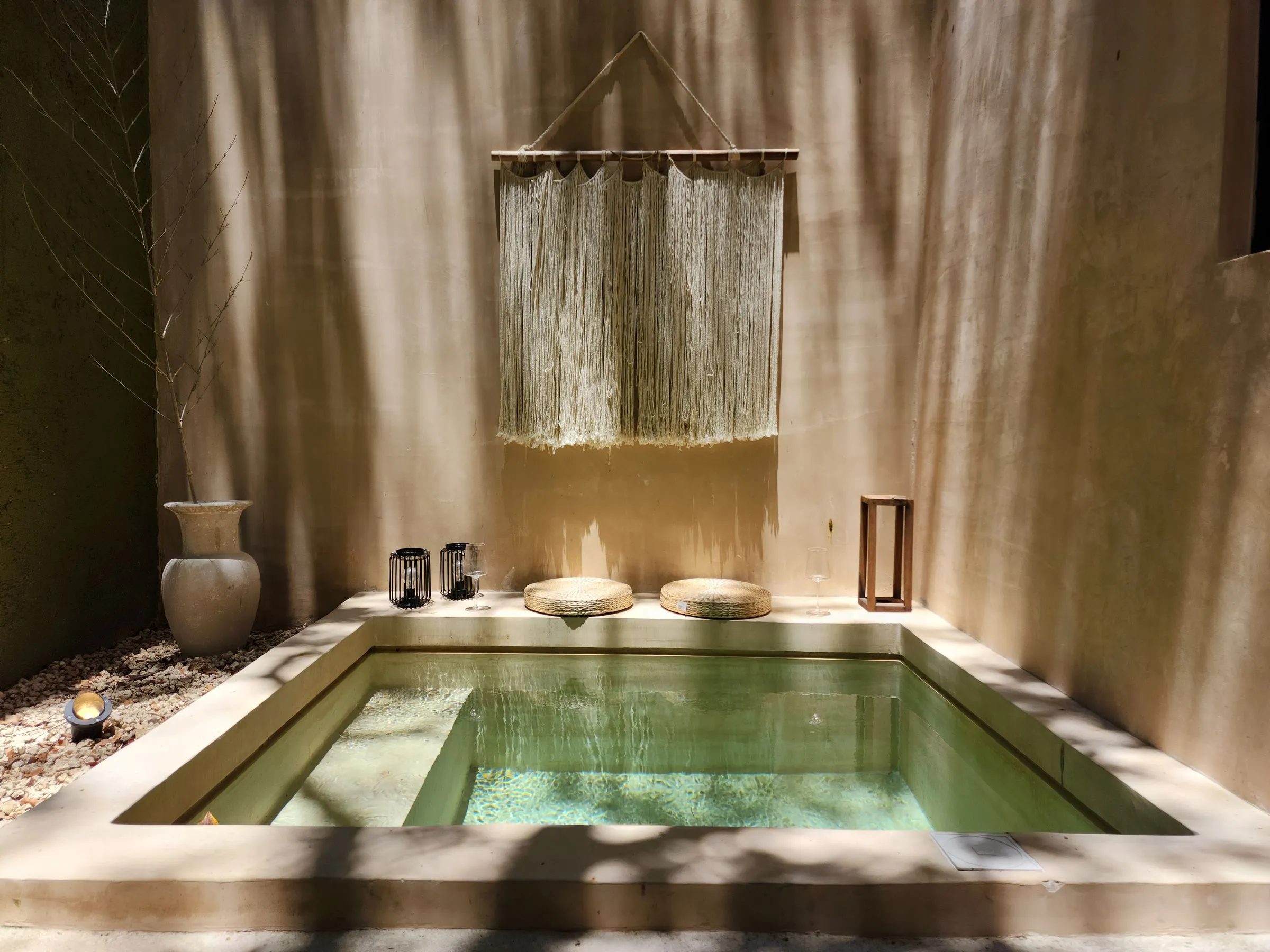 A sunlit stone soaking pool in a quiet walled courtyard, with woven cushions along its edge